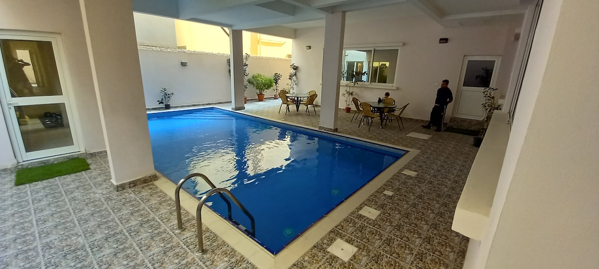 A serene pool area is featured, surrounded by a tiled floor and lush potted plants. The clear blue water of the pool is complemented by a set of lounge chairs and a dining table with chairs arranged nearby, creating a welcoming space for relaxation.