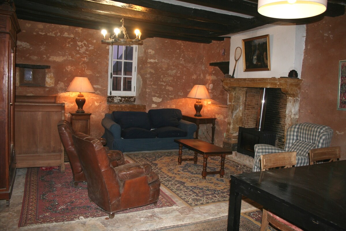 A warm living area features rustic exposed walls and a traditional fireplace. Soft seating options include an armchair and a sofa, complemented by a wooden coffee table. Two lampstands provide ambient lighting, while a patterned rug adds texture to the floor.