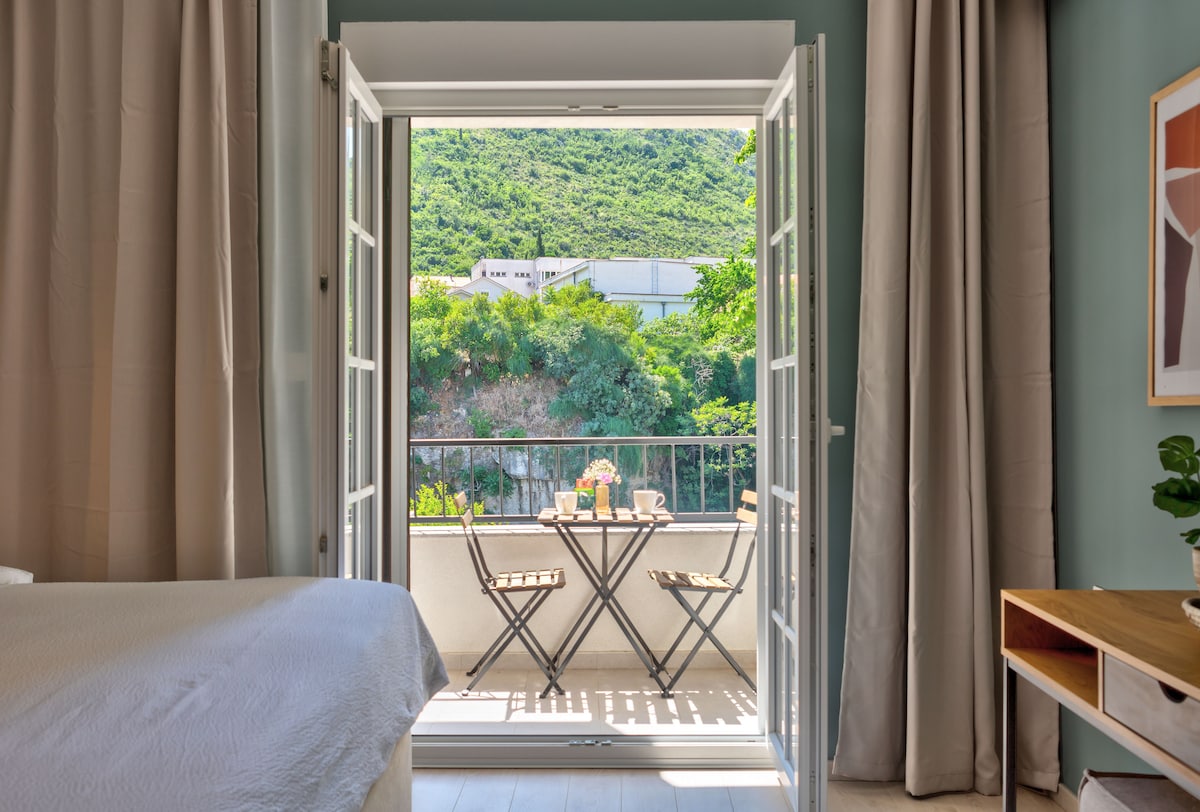 French doors open to a small terrace featuring two metal chairs and a circular table. Lush greenery is visible beyond the railing, complemented by mountain scenery. Soft curtains frame the doors, while a cozy bed and wooden furniture are arranged in the interior space.