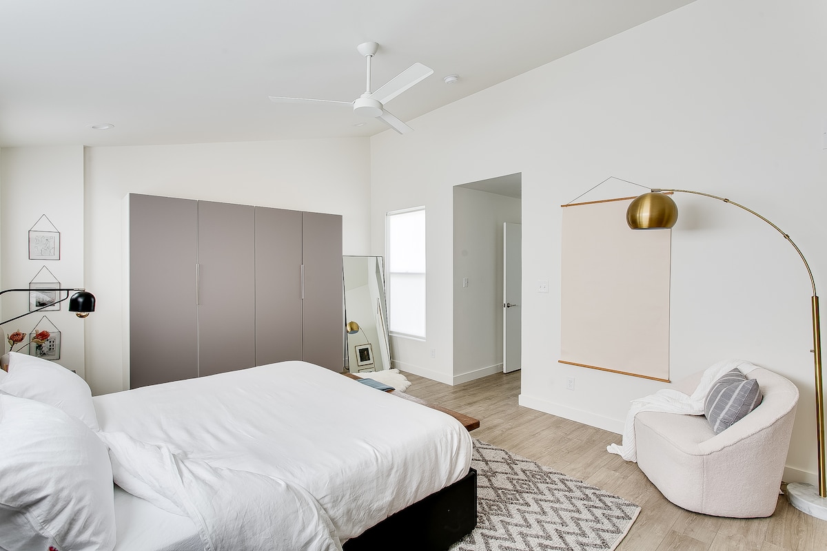 A spacious bedroom features a large bed with crisp white linens and a light-colored accent chair. A full-length mirror reflects natural light, and a modern floor lamp adds a touch of elegance. A closet with sliding doors offers ample storage.