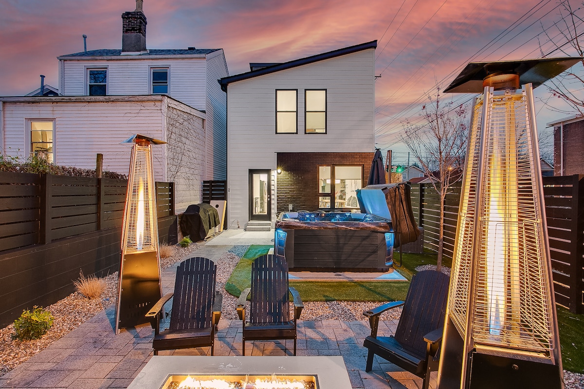 A backyard is displayed, featuring a hot tub and a gas fire pit surrounded by four outdoor chairs. Patio heaters provide warmth, and the space is framed by a fence. The modern home is visible in the background, illuminated by evening light.