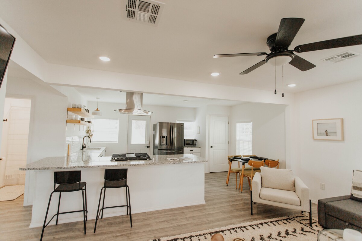 An open concept living space is presented, featuring a kitchen with a granite countertop and modern appliances. Bar stools are arranged at the counter, and a dining area is visible nearby. Natural light streams through multiple windows, illuminating the minimalist decor and comfortable seating.