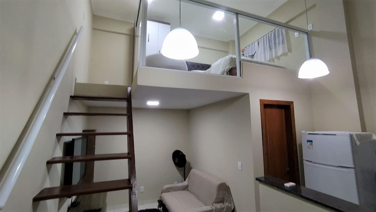 A loft space is shown, featuring a staircase leading to a mezzanine with a sleeping area. A light fixture hangs above the living area, which includes a sofa and a television mounted on the wall. A refrigerator is visible in the background.