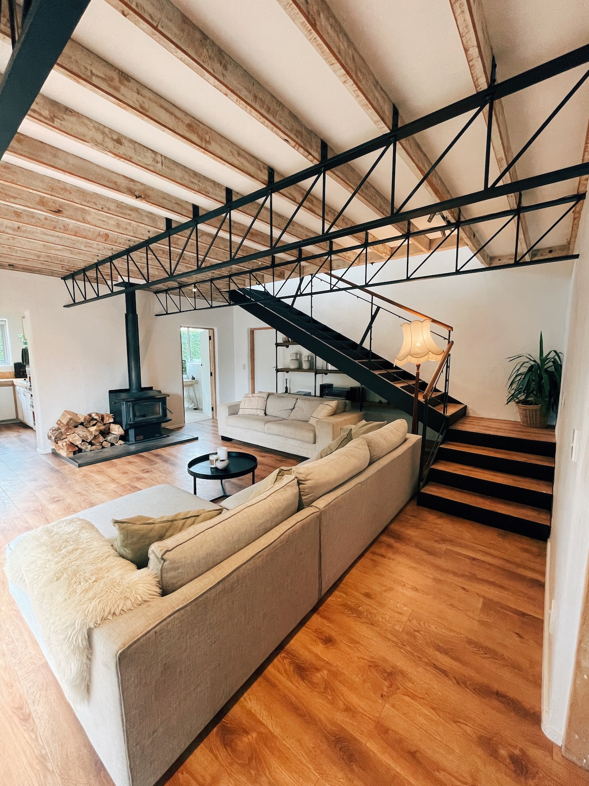 An open living area features a spacious sofa, a coffee table, and a cozy wood stove. Above, exposed wooden beams add character to the ceiling. A staircase with a modern railing leads to the upper level, while natural light fills the room through nearby windows.