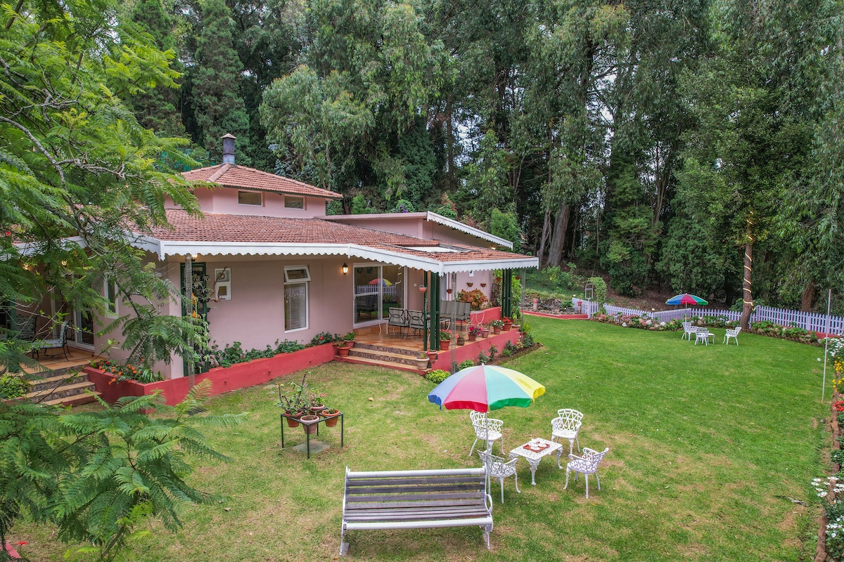 A charming bungalow sits amidst lush greenery, surrounded by well-maintained gardens. Colorful umbrellas shade outdoor seating areas, while a wooden bench adds to the inviting atmosphere. The spacious terrace is accessible, providing views of the peaceful surroundings and inviting relaxation.