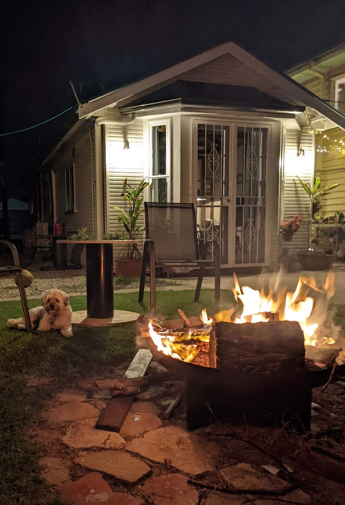 A fire pit radiates warmth in the foreground, surrounded by logs and stones. In the background, the cottage features decorative lighting and double doors framed by greenery. A dog rests peacefully by the fire, adding to the cozy evening ambiance.