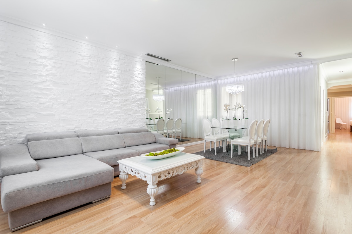 A light-filled living area features a spacious grey sectional sofa positioned against a textured white wall. A glass dining table with white chairs is visible in the background, separated by sheer curtains. Minimalist decor and natural wood flooring create a modern ambiance.