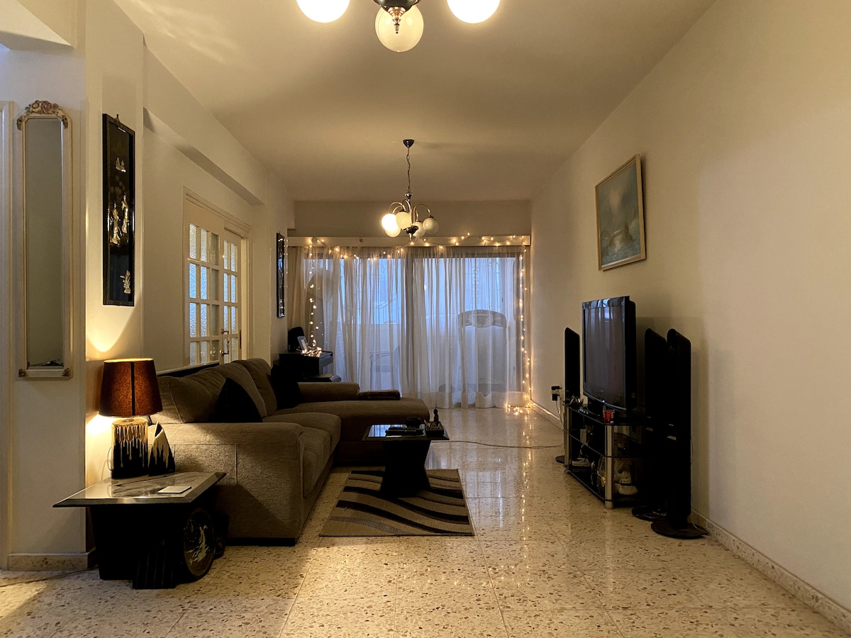 An airy living room showcases a spacious grey sofa paired with a dark coffee table. Soft lighting illuminates the area, highlighted by decorative string lights along the windows. A television stands against the opposite wall, with light curtains allowing natural light to filter through.