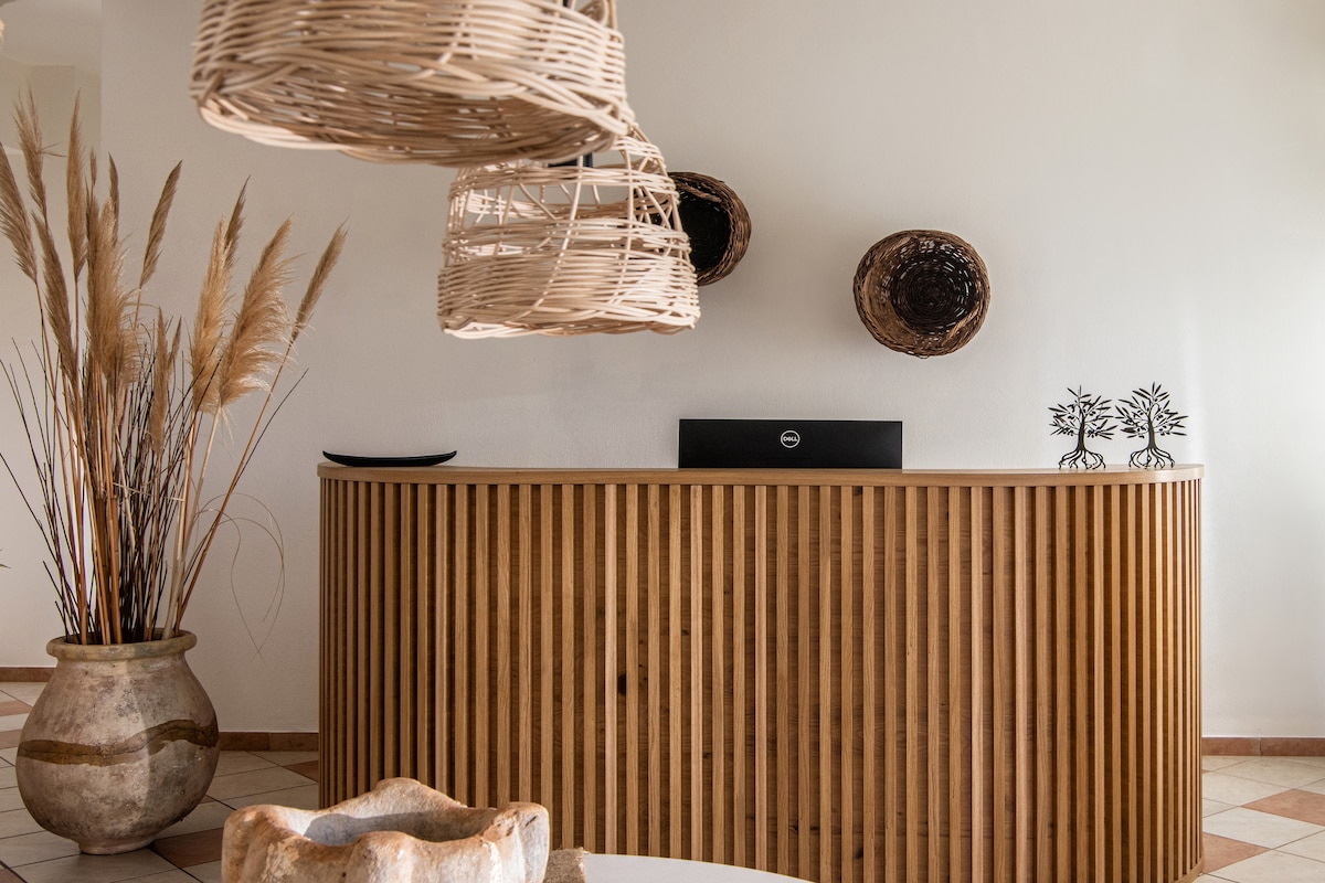 A reception area features a sleek wooden counter with vertical slats, complemented by natural woven light fixtures. Decorative wall baskets add texture to the space, while tall pampas grass is arranged in a ceramic vase, creating a welcoming atmosphere.
