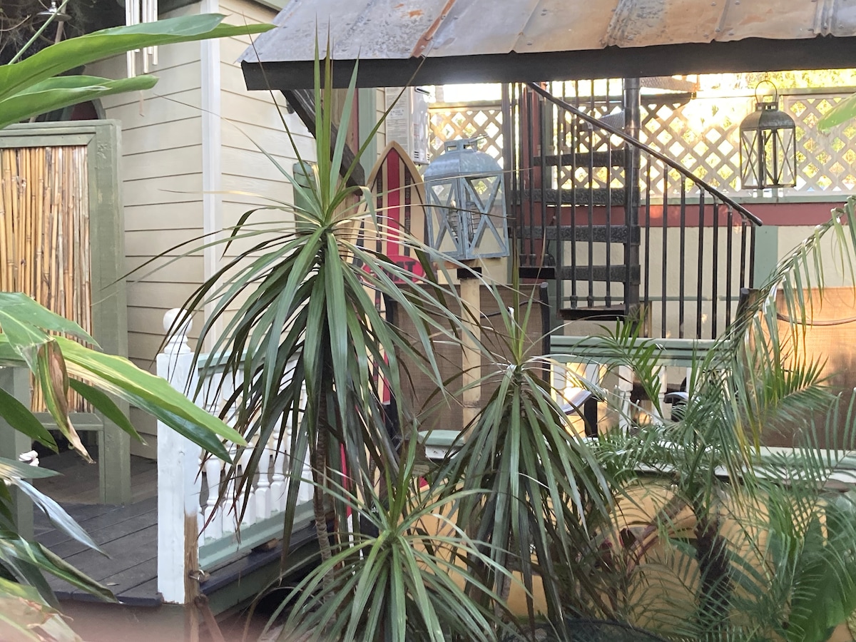 Lush tropical plants surround a wooden deck area, providing a natural backdrop. A staircase leads upward to the Treehouse, while lanterns and vibrant decor elements enhance the inviting atmosphere. The design reflects a blend of compact living and nature.