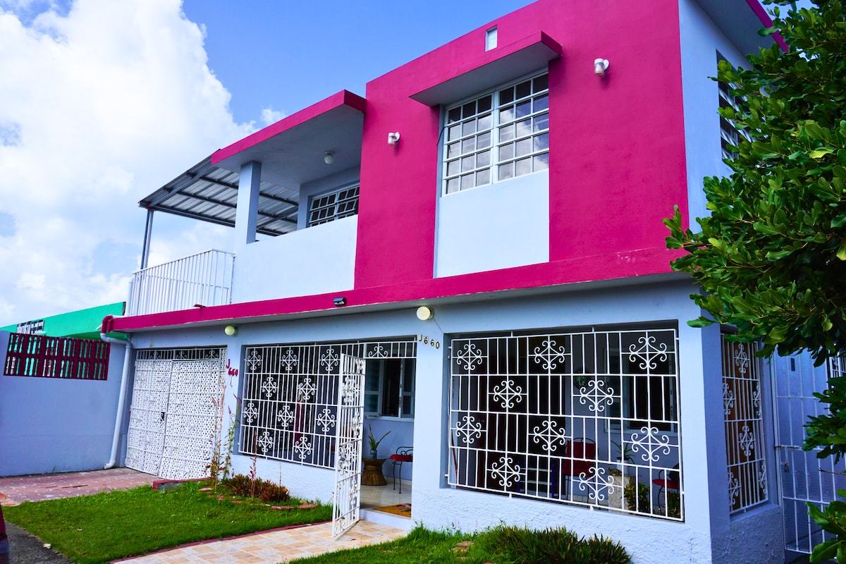 A freshly remodeled house features a vibrant pink upper facade with large windows surrounded by decorative wrought iron. An inviting entrance showcases a manicured lawn with small shrubs, while a covered terrace provides outdoor space. Ample parking is accessible in the front area.
