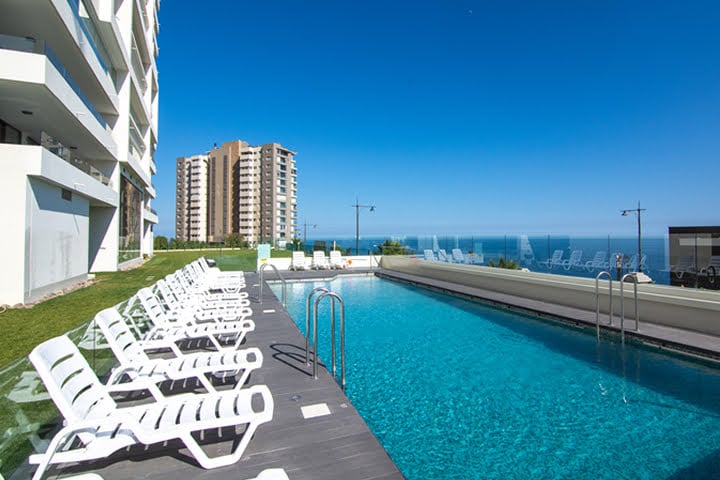 An outdoor pool area is featured, surrounded by comfortable lounge chairs arranged neatly along a deck. The serene blue water reflects the clear sky, with distant ocean views visible beyond the pool's edge. Nearby, modern buildings stand, contributing to the tranquil atmosphere.