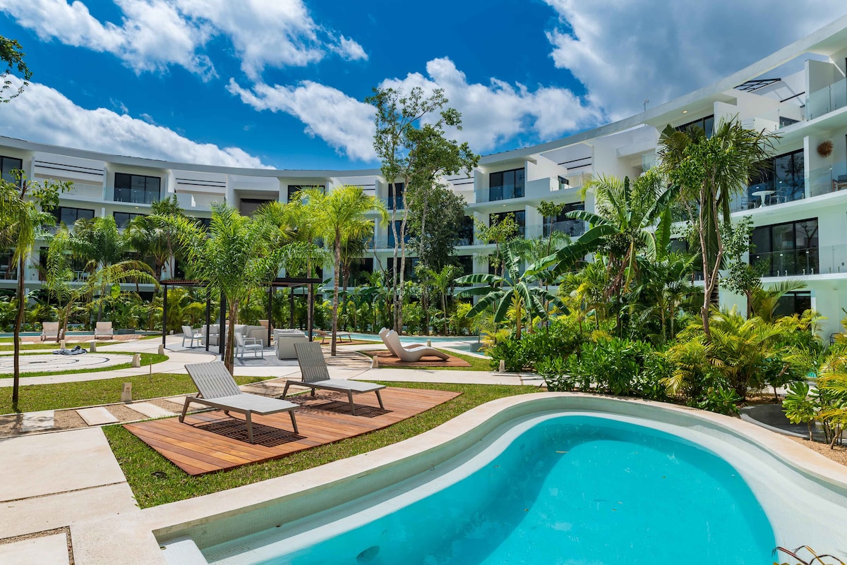 The image presents a lush courtyard featuring multiple manicured gardens, tropical plants, and modern architecture. Several lounge chairs are positioned near a serene pool, while pathways meander through the space. The clear blue sky enhances the inviting atmosphere of this tranquil environment.