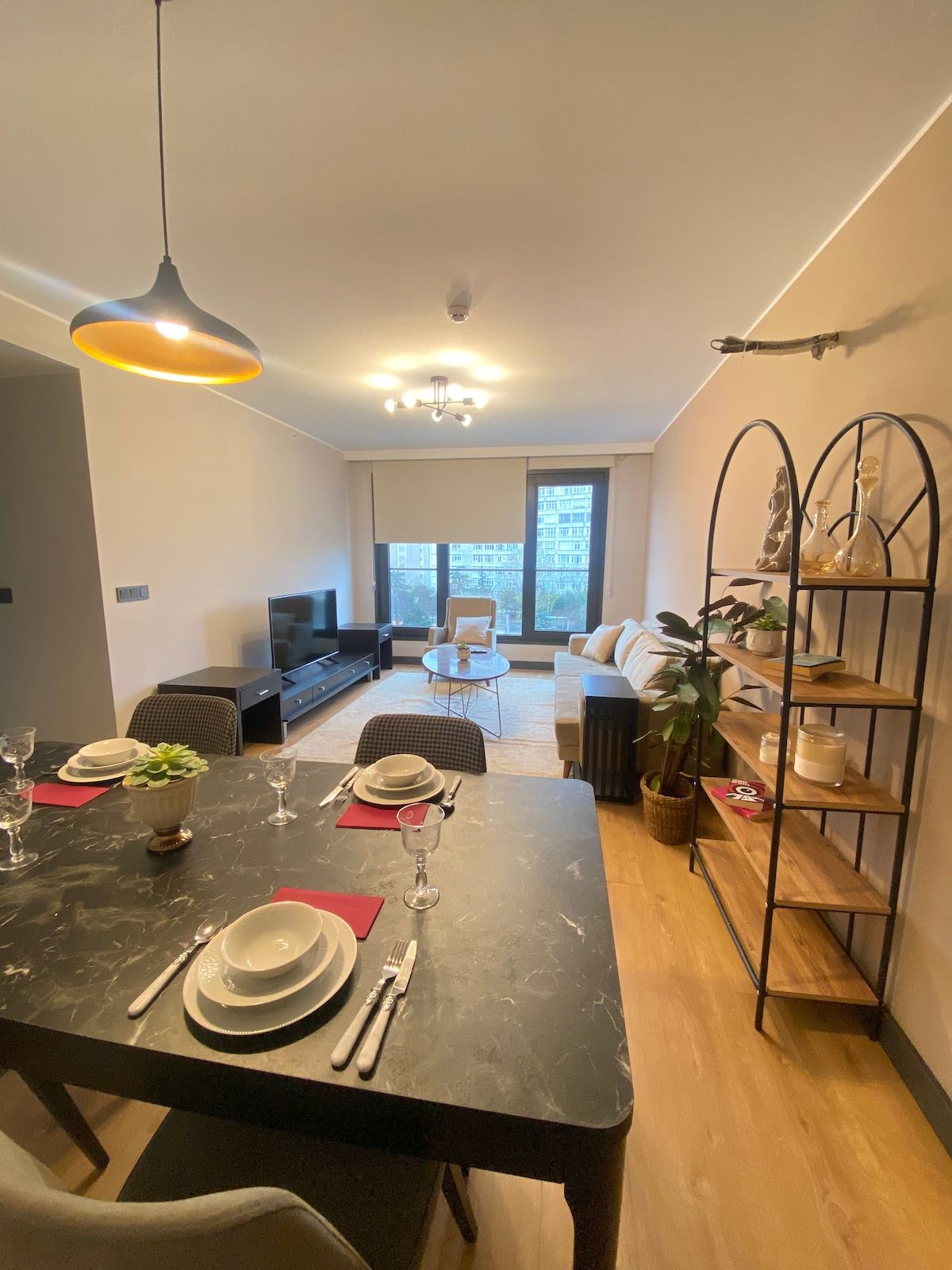 A modern living space features a dining area with a dark table set for four. A comfortable sofa and a single chair are arranged near large windows, providing natural light. Decorative plants and a stylish shelving unit enhance the room's ambiance.