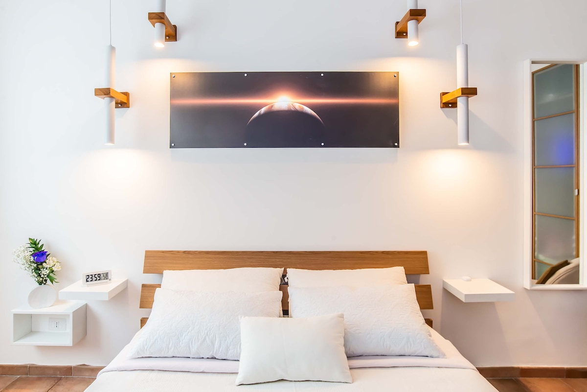 The bedroom features a wooden bed with light-colored bedding arranged neatly. Above the bed, a wall art piece depicts a celestial scene, illuminated by two wall-mounted lights. Small shelves on either side of the bed offer decorative items, enhancing the serene ambiance.