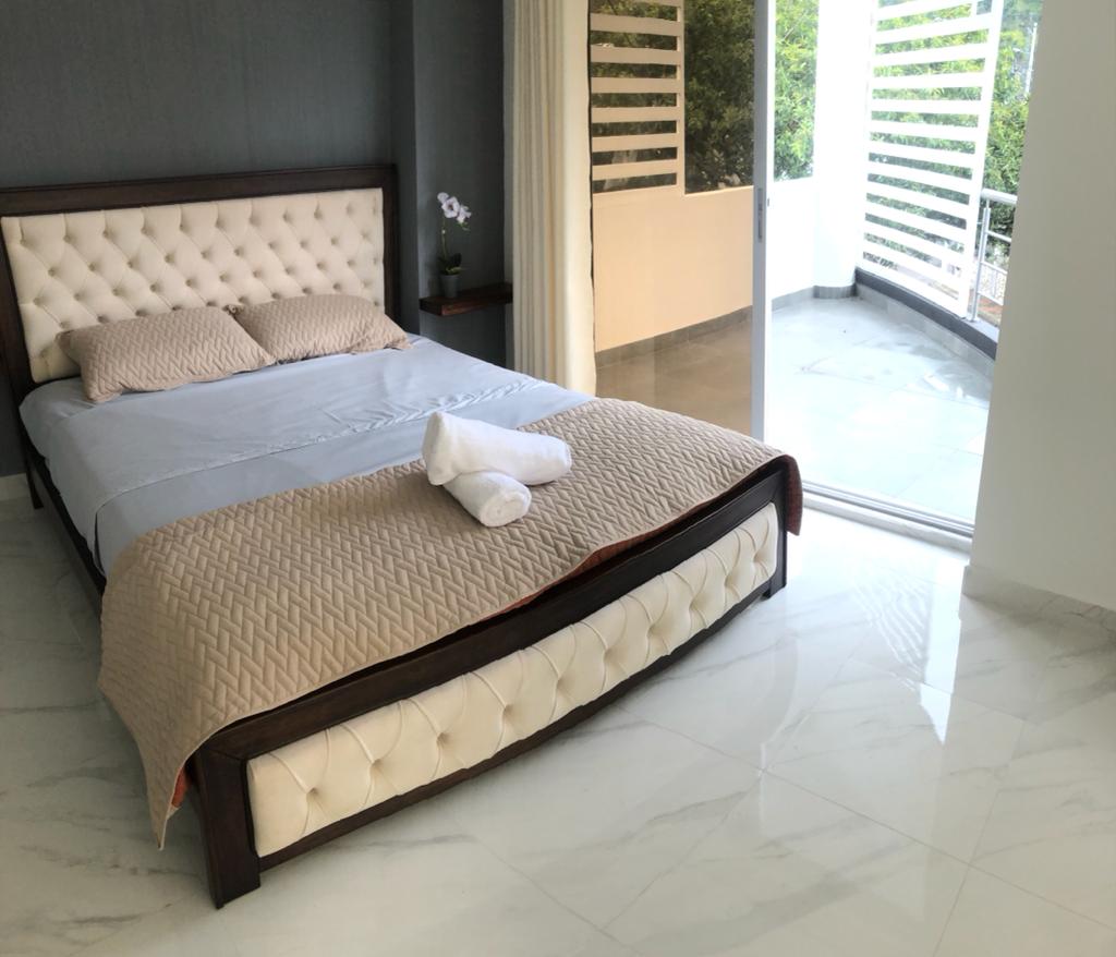 A spacious bedroom features a tufted bed with a light-colored blanket draped neatly. Fresh towels are positioned on the bed, and large glass doors lead to a private balcony, allowing natural light to brighten the room.