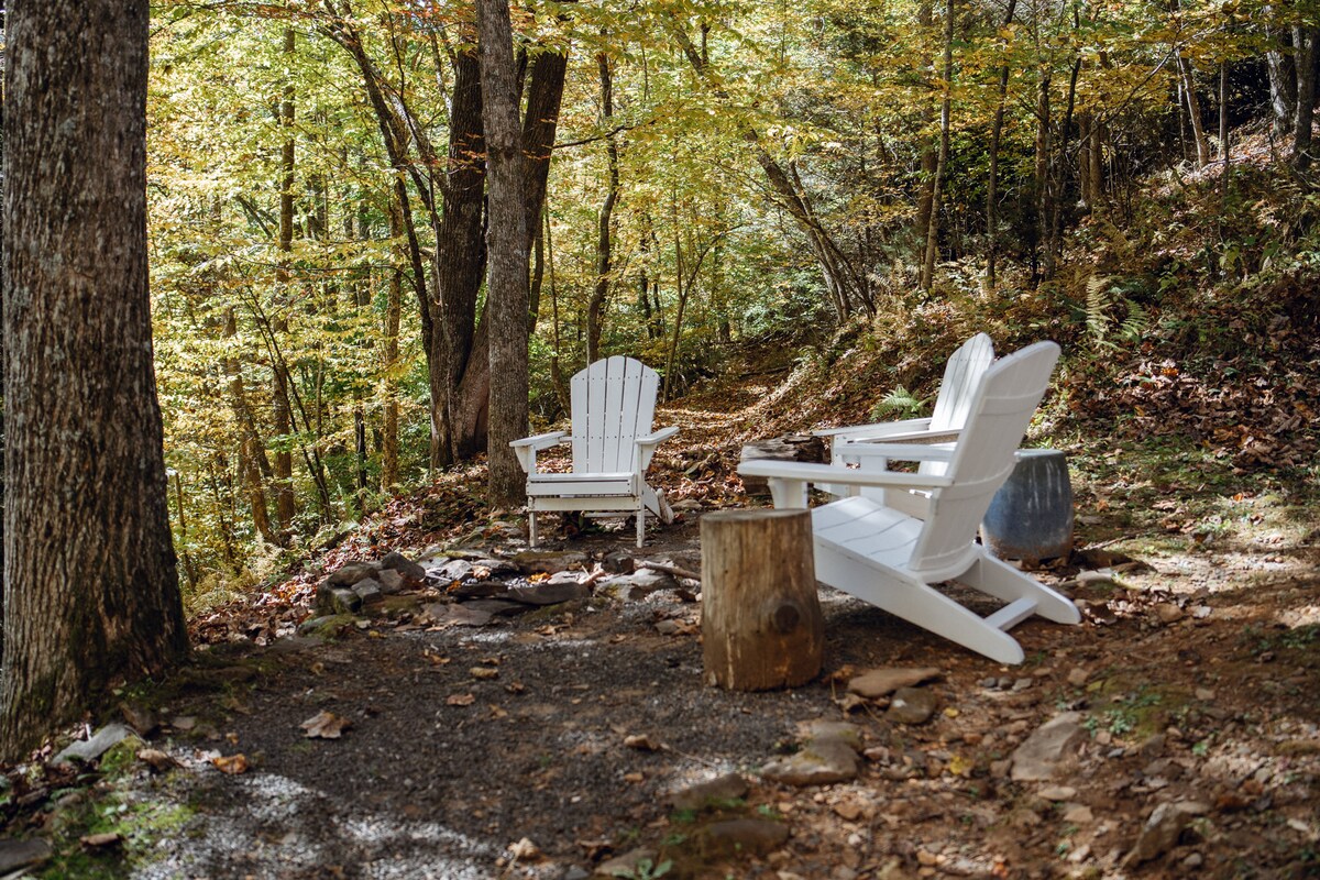 Two white Adirondack chairs are positioned in a natural clearing, surrounded by trees with golden leaves. A small log serves as a rustic side table, and a stone and gravel path leads through the serene outdoor setting.