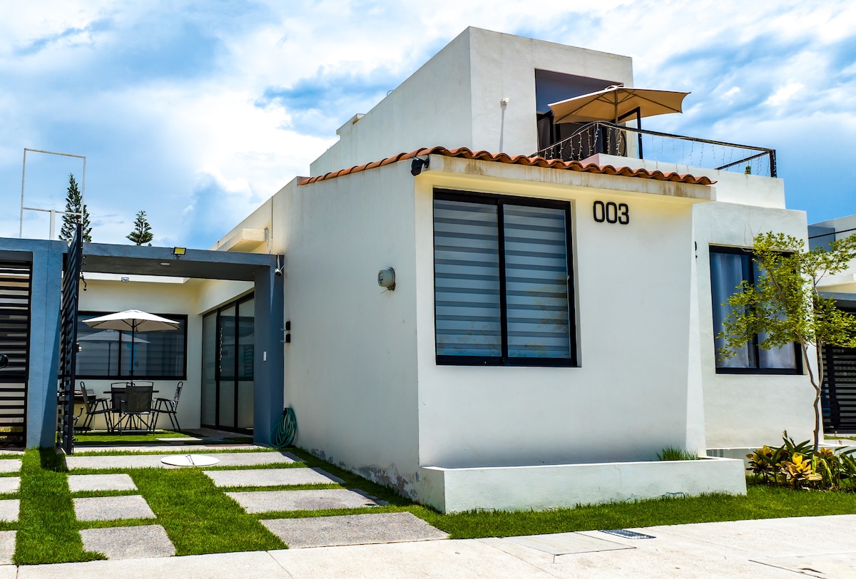 A modern house is presented with a clean white exterior and a terracotta-hued roof. Large windows allow natural light to enhance the interior. The welcoming entrance is accompanied by a well-maintained garden featuring stone pathways and outdoor seating under an umbrella.