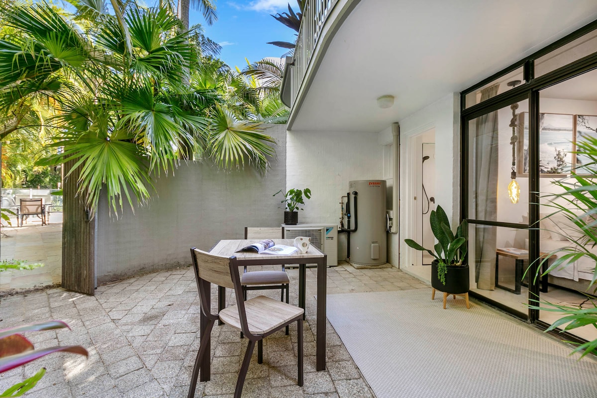 A private outdoor patio is surrounded by lush greenery, featuring a small table and two chairs. A water dispenser is positioned near the entrance of the unit, while textured tiles and a soft rug add to the inviting atmosphere.