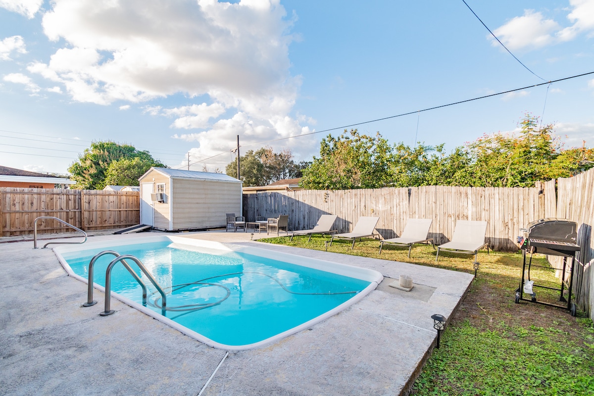 An outdoor space features a sparkling inground pool surrounded by a concrete deck. Several lounge chairs are positioned nearby, offering relaxation by the water. The fenced area is bordered by greenery and a grill is placed strategically for outdoor cooking.