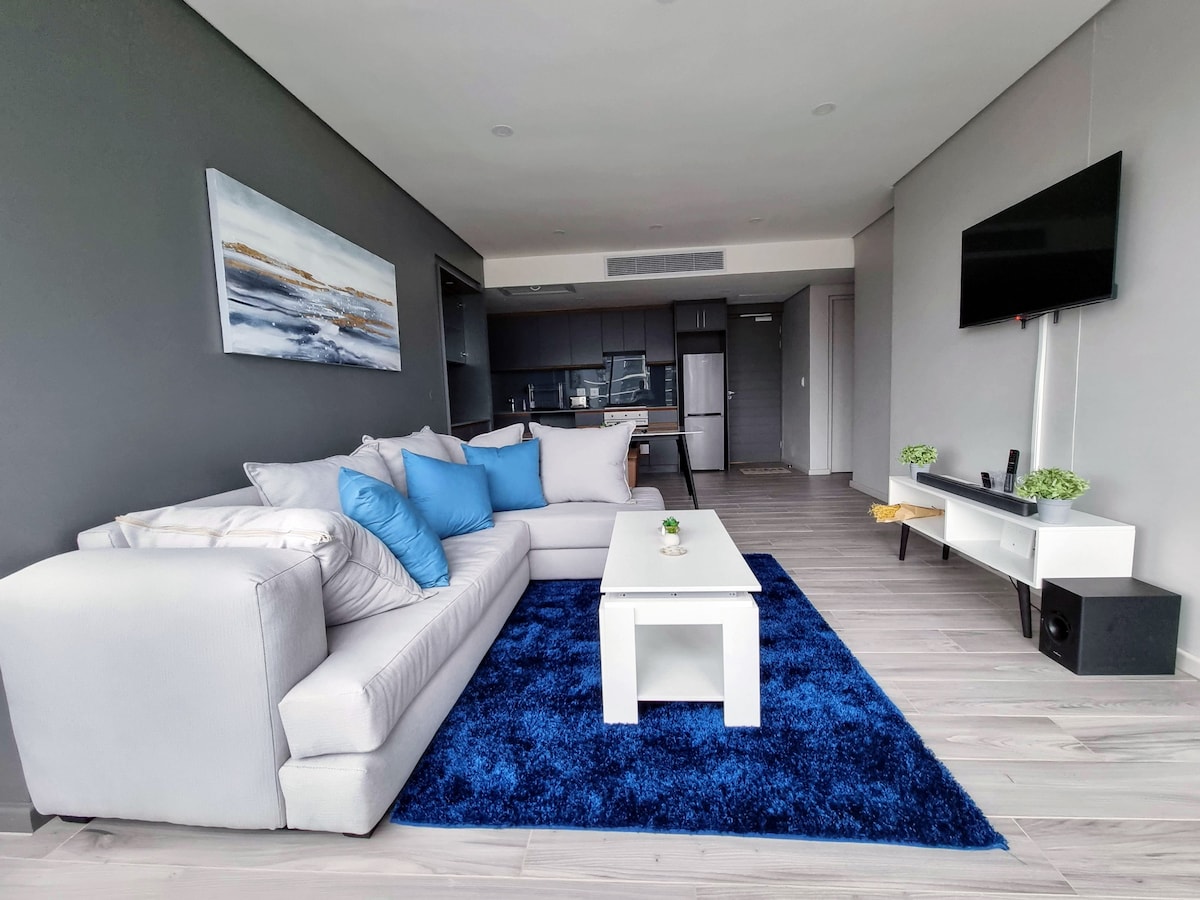 A modern lounge area is furnished with a light-colored sectional sofa adorned with blue cushions. A white coffee table is positioned on a deep blue area rug, complementing the space. A wall-mounted TV is visible, and the kitchen area is seen in the background.