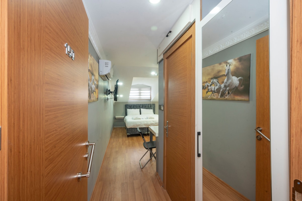 A hallway is shown featuring wooden doors leading to rooms. A small table with a chair is positioned on one side, while soft lighting illuminates the space. Artwork depicting horses decorates the wall in the background, and textured flooring enhances the overall appearance.