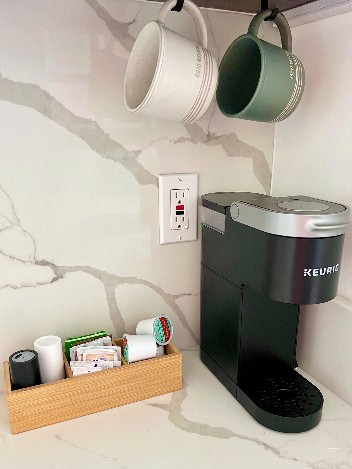 A coffee station is featured with a Keurig machine positioned against a marble backsplash. Two ceramic mugs, one white and one green, hang above. A wooden container holds various coffee pods and supplies, providing convenience for morning routines.