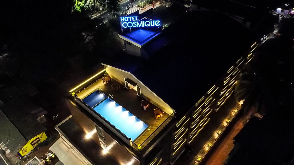 An aerial view of Hotel Cosique is displayed, showcasing a rooftop swimming pool illuminated by soft lighting. The hotel's modern architecture features multiple floors and a rooftop terrace lined with lounge chairs. The hotel name is prominently displayed, adding a touch of character to the urban skyline.