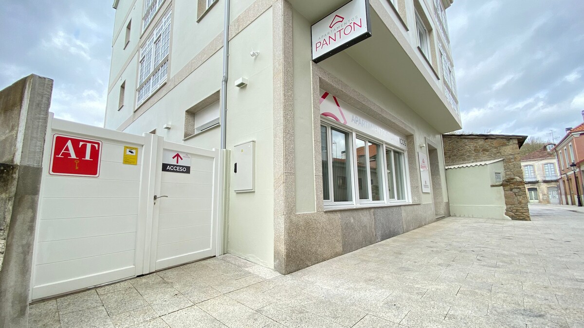 The exterior of the building presents a modern design with large windows allowing natural light to enter. A white gate provides access to the property, and signage is visible indicating the building's name. The surrounding area features paved pathways and a clean, organized appearance.