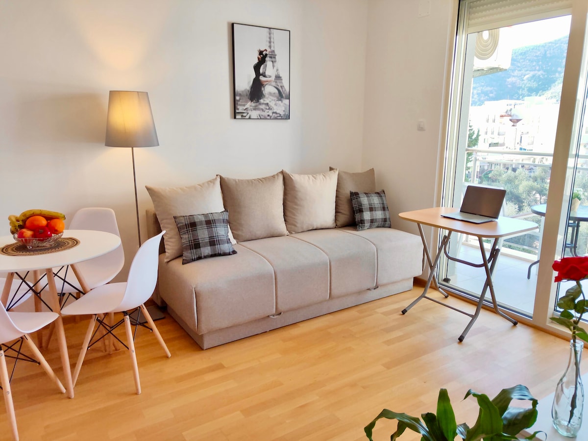 A cozy living space features a comfortable sofa with decorative pillows, complemented by a small dining table with white chairs. A lamp adds soft illumination, and a laptop is positioned on a foldable table. Large glass doors provide access to a balcony with a scenic view.