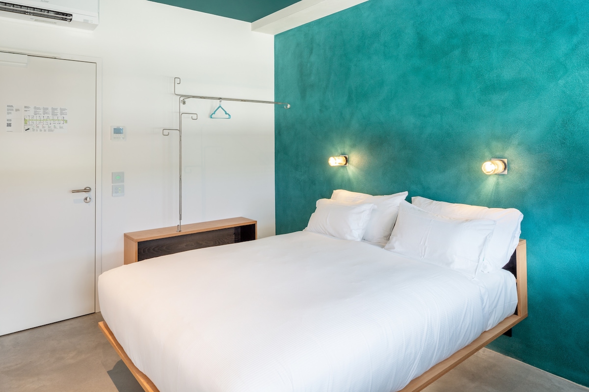 A modern bedroom features a spacious bed against a vibrant turquoise wall. Stylish wall-mounted lighting provides soft illumination. A minimalist wooden dresser and a simple clothes rack are positioned nearby, contributing to the clean and functional design of the space.