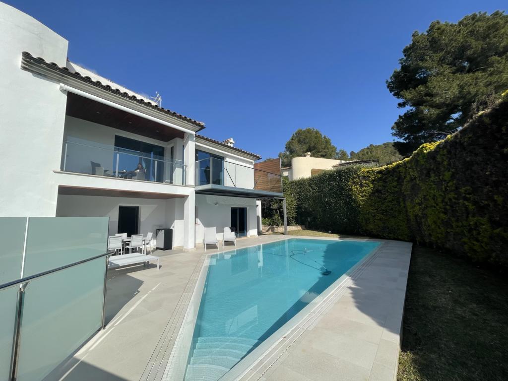 The exterior view of the villa features a modern design with large windows and a spacious terrace. A tranquil pool is situated in front, surrounded by well-maintained greenery. Sun loungers are positioned beside the pool, creating a serene outdoor living space.