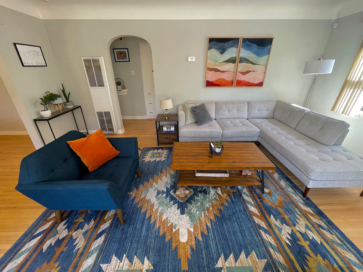 A spacious living area is featured with a gray sectional sofa and a blue accent chair, complemented by a wooden coffee table. An area rug with a colorful design covers the floor. Two pieces of abstract wall art adorn the wall, while natural light enters through a window.