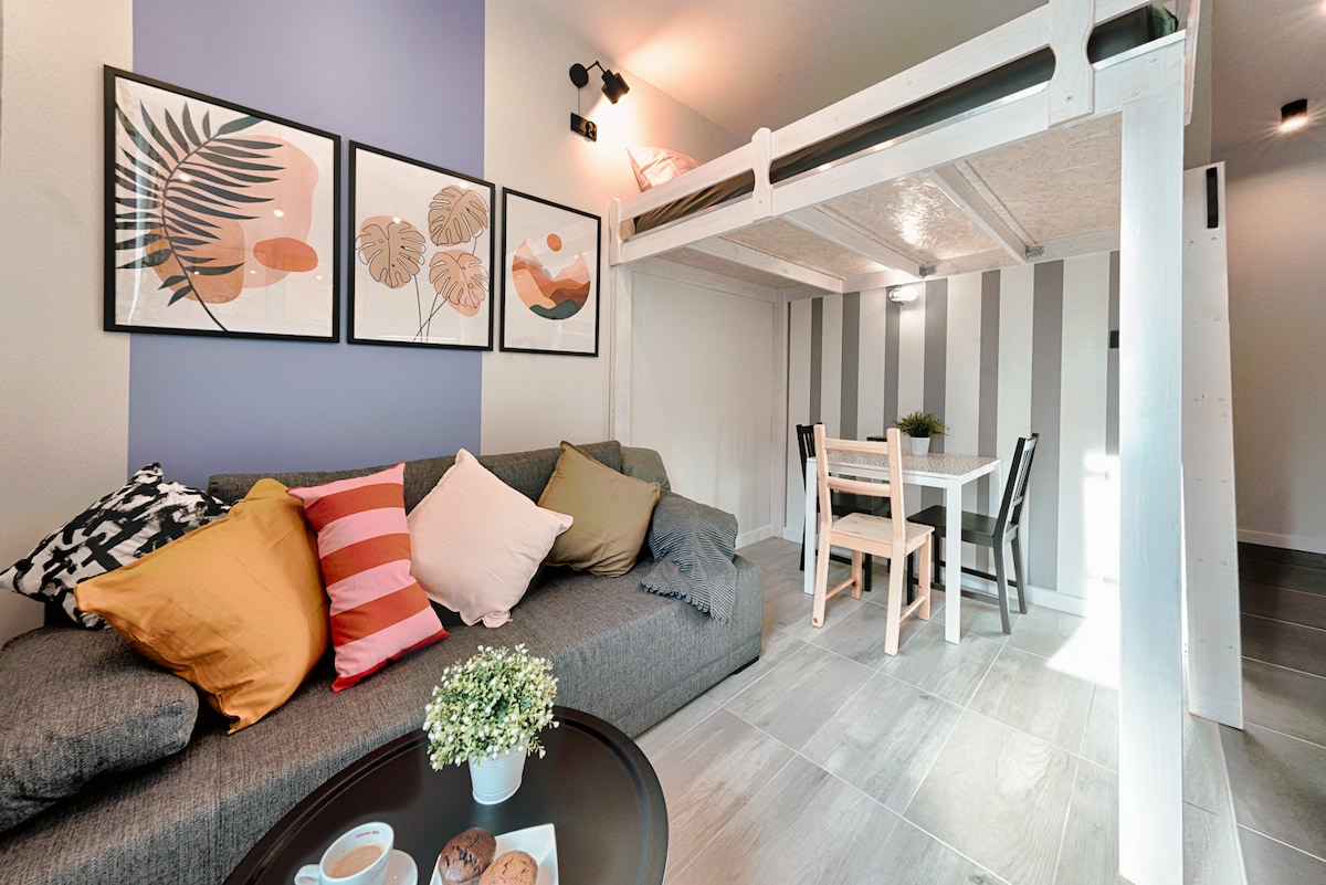 A studio apartment features a cozy seating area with a sectional sofa adorned with various cushions. Above, a sleeping loft is visible. A dining table with chairs is situated nearby, complemented by framed artwork on the wall showcasing botanical themes.