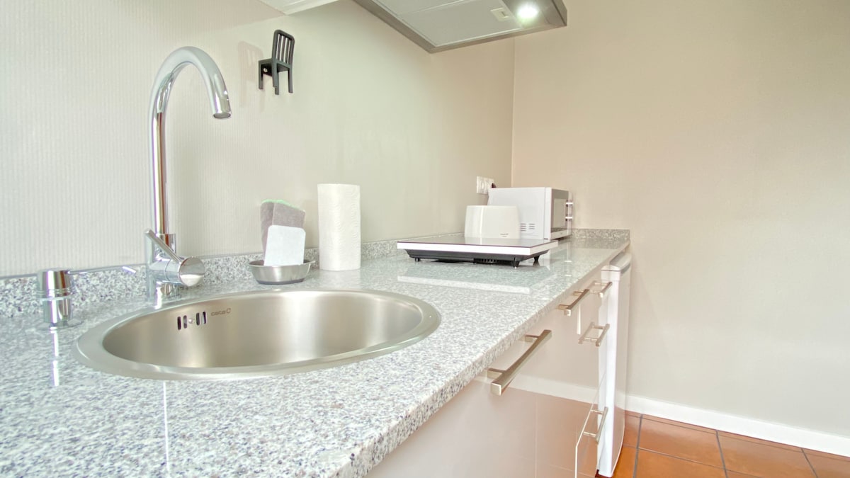 A modern kitchen area is presented with a sleek countertop and stainless-steel sink. A microwave and other kitchen appliances are neatly arranged, highlighting the convenience of the space. Under-cabinet lighting adds visibility to the area, enhancing functionality.