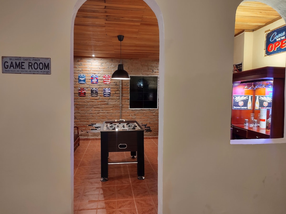 The game room is accessible through an arched entrance, featuring a foosball table at the center. Brick walls add a rustic touch, while posters are displayed above. A flat-screen television is mounted on one wall, and ambient lighting is provided by a pendant lamp hanging overhead.