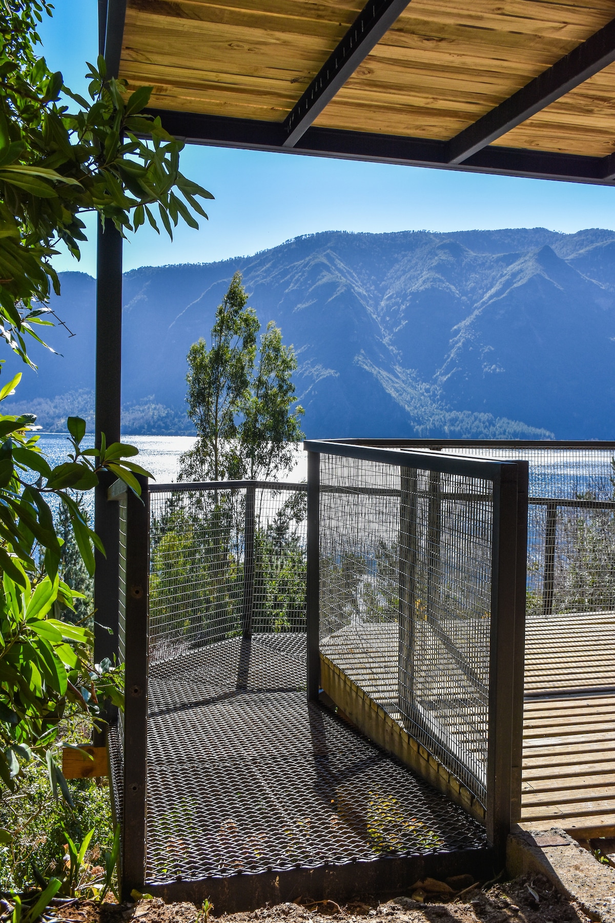 A spacious balcony features a metal railing with a mesh design, providing safety and a clear view of the surrounding mountains and lake. Natural foliage frames the terrace, enhancing the outdoor connection while allowing plenty of sunlight to filter through.