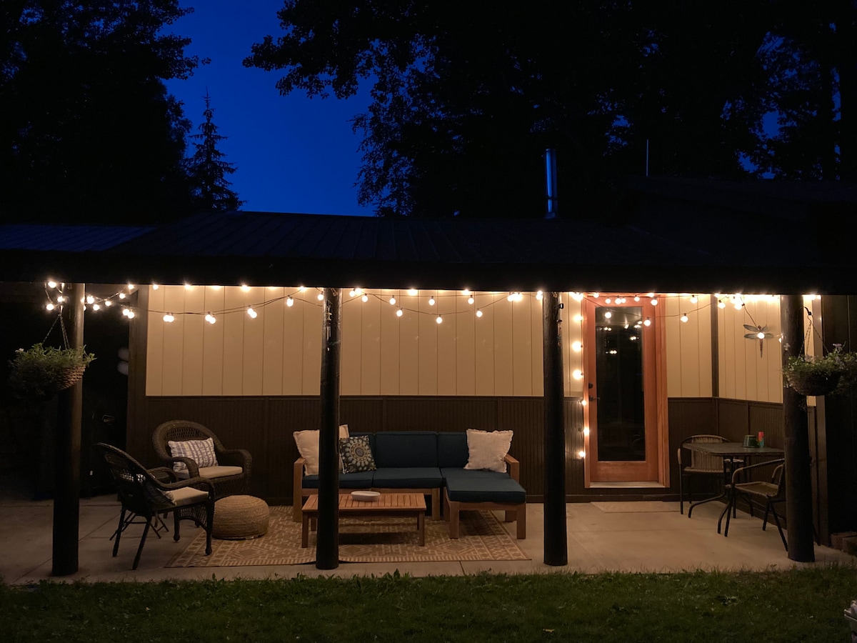 A covered patio area is illuminated by string lights, creating a warm ambiance. Comfortable seating is arranged around a wooden coffee table, while a small side table is also visible. The surrounding greenery enhances the tranquil setting, inviting relaxation outdoors.