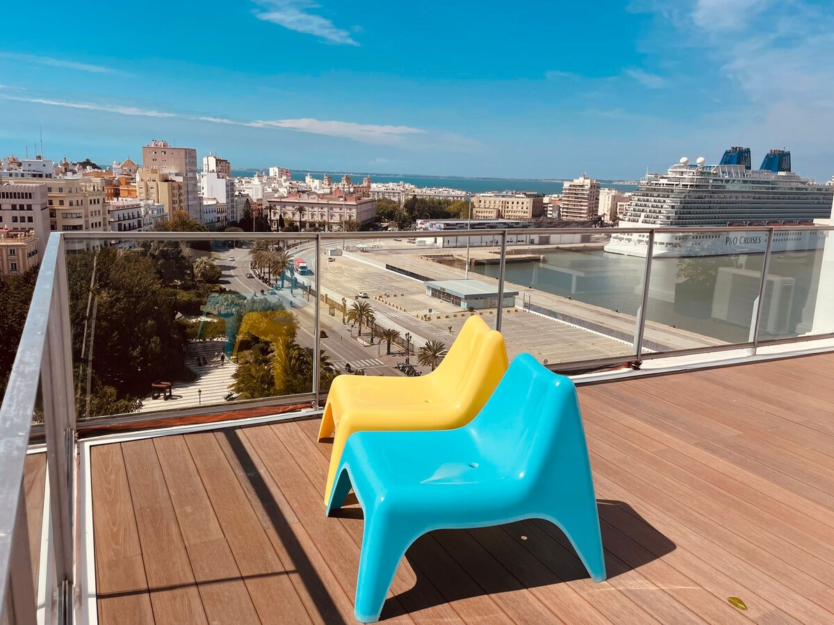 Two colorful lounge chairs, one yellow and one blue, are placed on a wooden deck overlooking a panoramic view of the city of Cádiz and the sea. The background features urban architecture and a harbor, with a clear blue sky above.