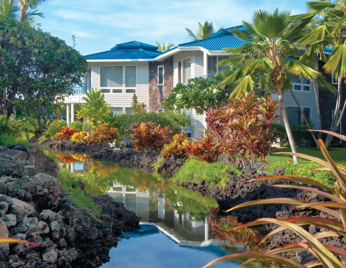The exterior of a tropical resort suite is depicted, featuring a blue-roofed building surrounded by lush greenery and colorful plants. A tranquil water feature reflects the building and foliage, enhancing the serene ambiance of the environment.