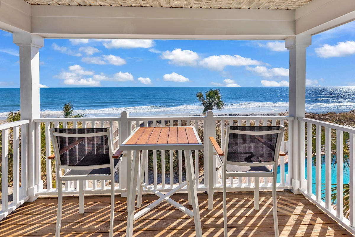 A serene outdoor balcony is showcased, featuring a table and two chairs. The bright ocean and sky extend beyond the railing, offering an unobstructed view of the coastline. Palms can be seen in the foreground, adding to the tranquil beach setting.