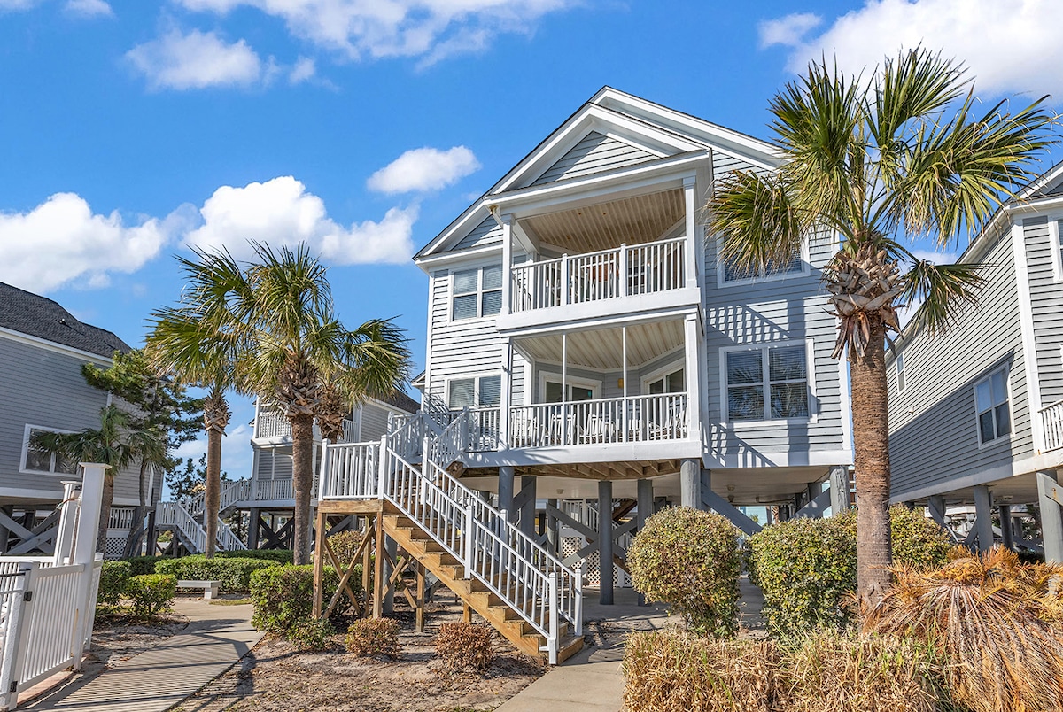 A two-story beach house is showcased, featuring a welcoming staircase leading to a spacious front porch. Surrounded by well-maintained palm trees and low shrubbery, large windows invite natural light into the home, highlighting its coastal architecture under a clear blue sky.