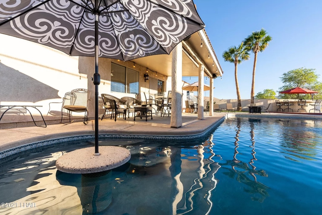 A relaxing outdoor area features a swimming pool bordered by paved stone. A patio umbrella with a black and white design provides shade over a table and chairs set. Palm trees are visible in the background, contributing to the serene atmosphere.