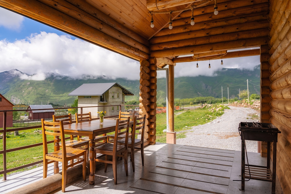 An outdoor terrace is furnished with a large wooden table and several chairs, providing a space for gatherings. The surrounding mountains are visible in the background, framed by a clear blue sky with hints of clouds. A small grill is positioned nearby.