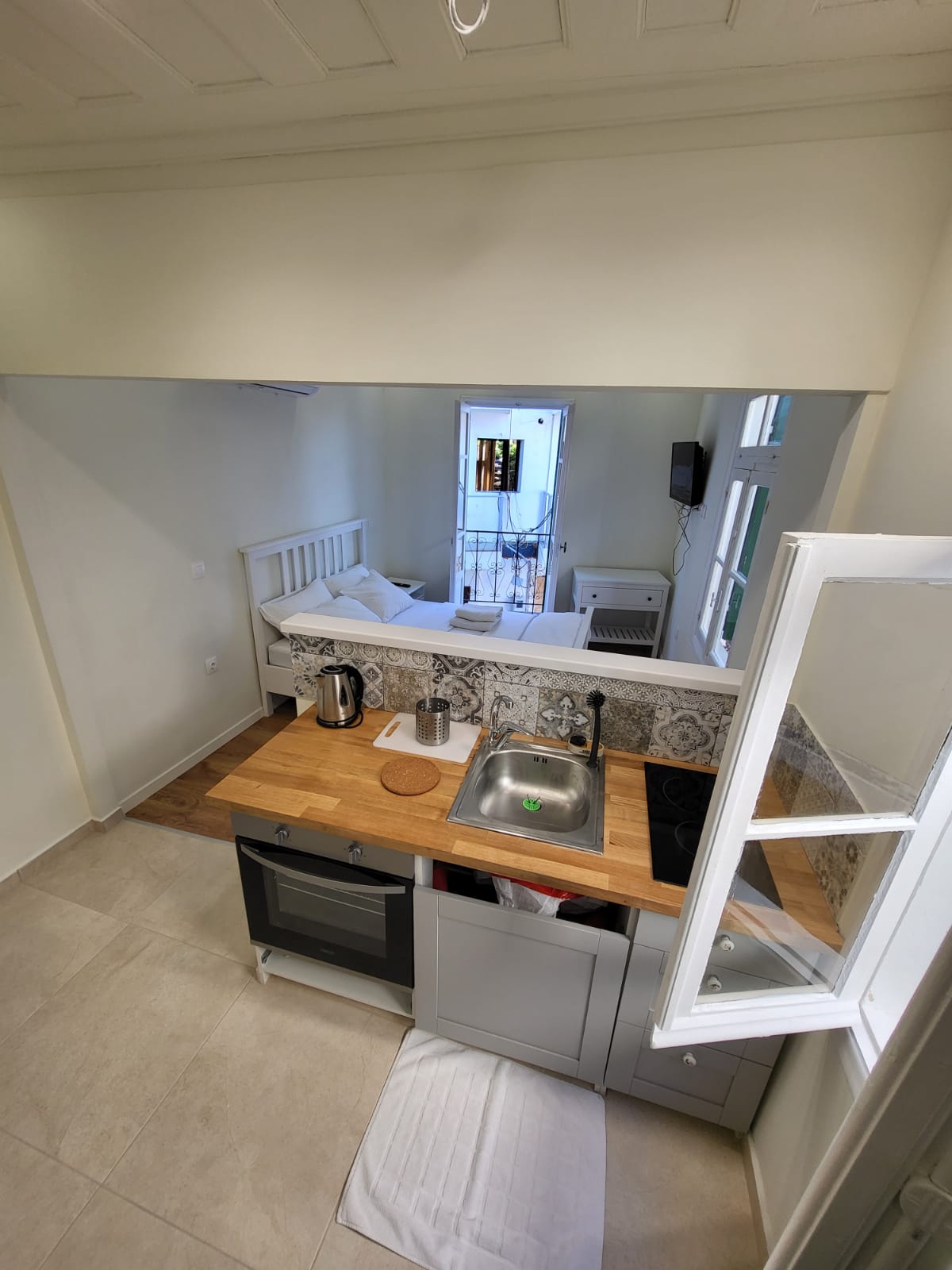 A modern kitchen area is presented with wooden countertops and gray cabinetry. A sink is visible alongside a kettle and cooking appliances. Natural light enters from a nearby window, illuminating the minimalist design. A glimpse of the sleeping area is seen through the open space.