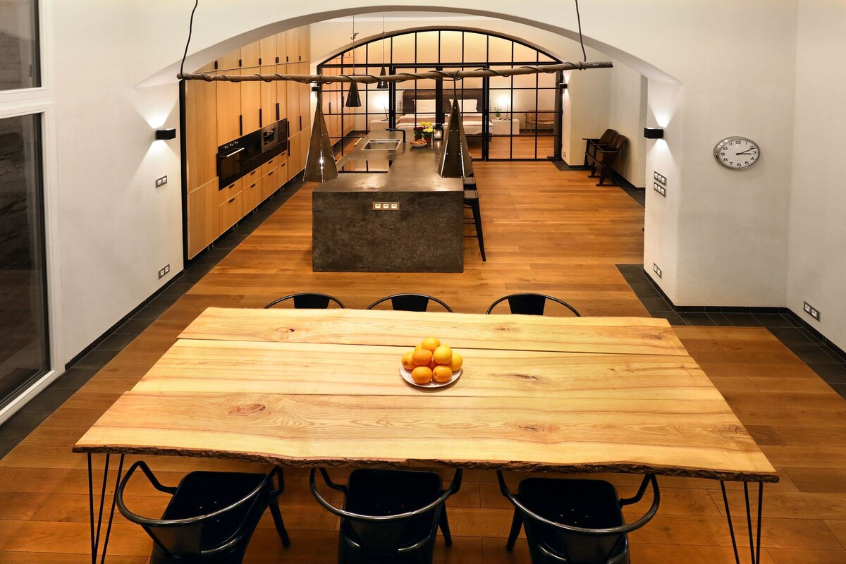 A spacious dining area features a large wooden table set with a bowl of oranges. Black chairs surround the table, with the kitchen viewable in the background. Natural light floods the room, highlighting the wood flooring and modern design elements.