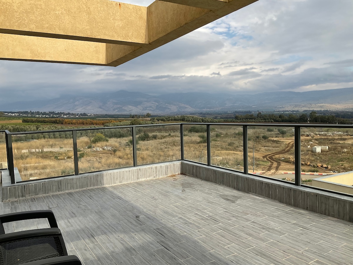A spacious balcony features a glass railing, providing an unobstructed view of the landscape. Rolling hills and distant mountains are visible under a cloudy sky. The balcony flooring is tiled, while two black chairs are positioned along the edge.