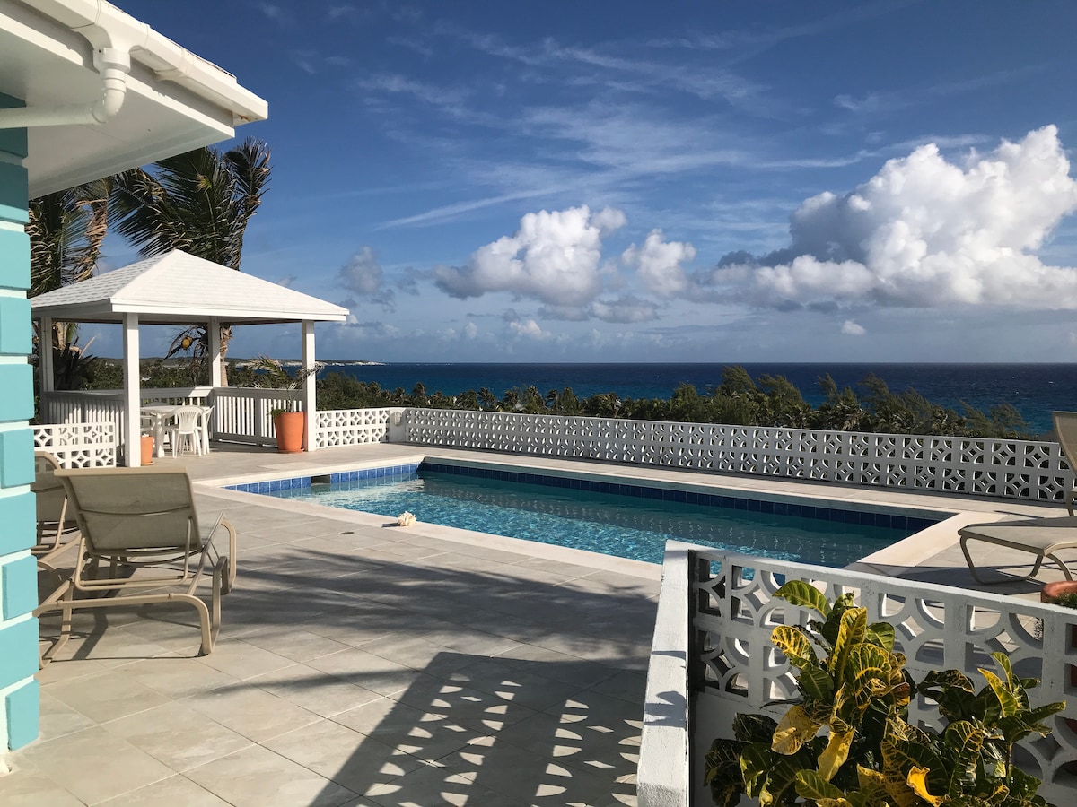 A bright pool terrace is visible, featuring a clear blue swimming pool surrounded by sun loungers. A gazebo with a white roof stands in the background, with lush greenery and the Atlantic Ocean beyond. Soft white clouds are scattered across a blue sky.