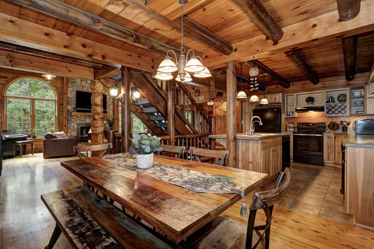 An open-concept living area features wooden beams and a spacious dining table made of natural wood, accompanied by metal chairs. The kitchen is visible in the background, showcasing modern appliances and warm lighting. Large windows provide ample natural light and views of the surrounding forest.