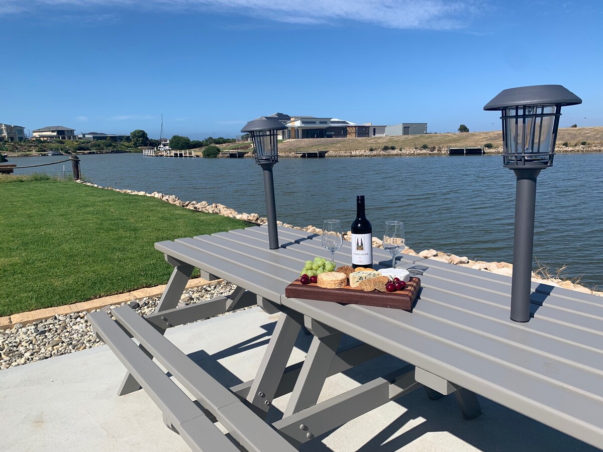 A picnic table is set beside the water, featuring a bottle of wine, glasses, and a platter with grapes and cheese. Two lanterns stand at the table, surrounded by green grass and a stone-lined edge, with a calm water view and buildings in the background.